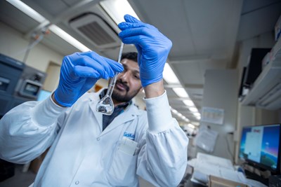 A SickKids staff member wearing a lab coat and blue gloves carefully pours a liquid from a pipette into a flask in a laboratory setting. He is focused on his task, with lab equipment and a computer screen visible in the background.